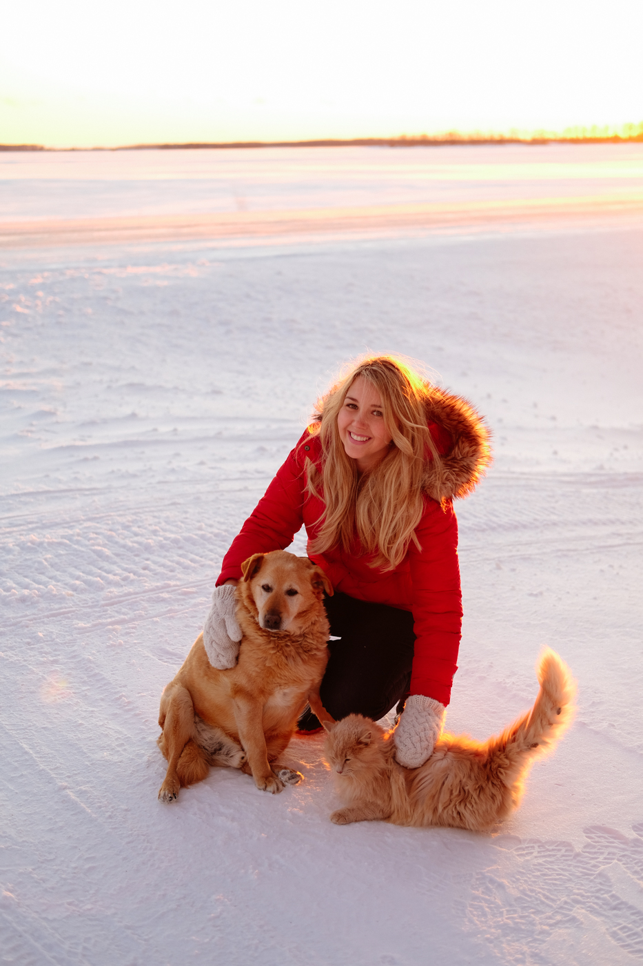 Debora Dahl wearing a red coat in the snow with the sunset behind her with a dog and a cat